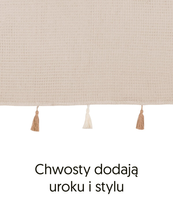 Brzeg poncza z bawełny waflowej w kolorze beżowym i ozdobne chwosty, z podpisem „Chwosty dodają uroku i stylu”.